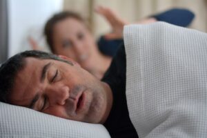Man asleep and snoring in bed with frustrated wife in background.